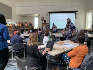 Man presenting to a crowd that is seated around multiple tables inside a library